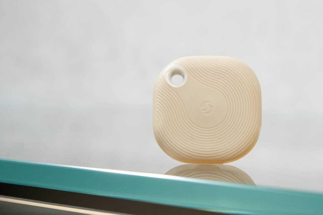 Un tracker beige, quadrato con angoli arrotondati e un foro per laccetto nell'angolo superiore, è poggiato su una superficie di vetro.