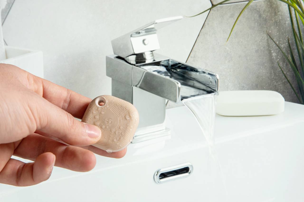 Una mano tiene un piccolo oggetto quadrato davanti a un rubinetto da cui scorre l'acqua. Sullo sfondo sono visibili delle piante.