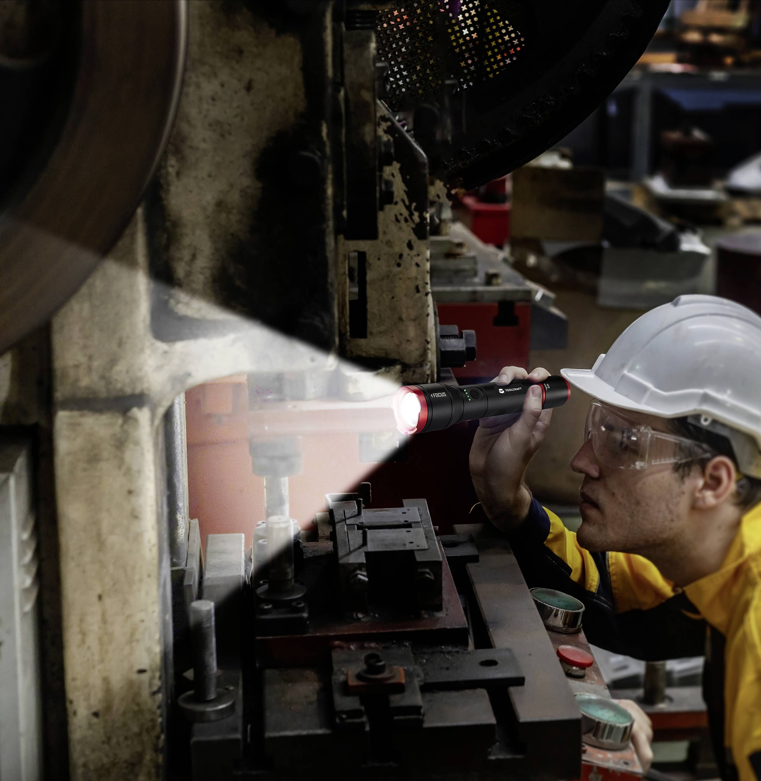 Un operaio con casco e occhiali di protezione ispeziona una macchina industriale utilizzando una torcia in un ambiente di fabbrica.