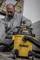 Una persona sta pulendo un cantiere con un aspirapolvere giallo DeWalt per uso umido e secco. Indossa abbigliamento da lavoro e guanti.
