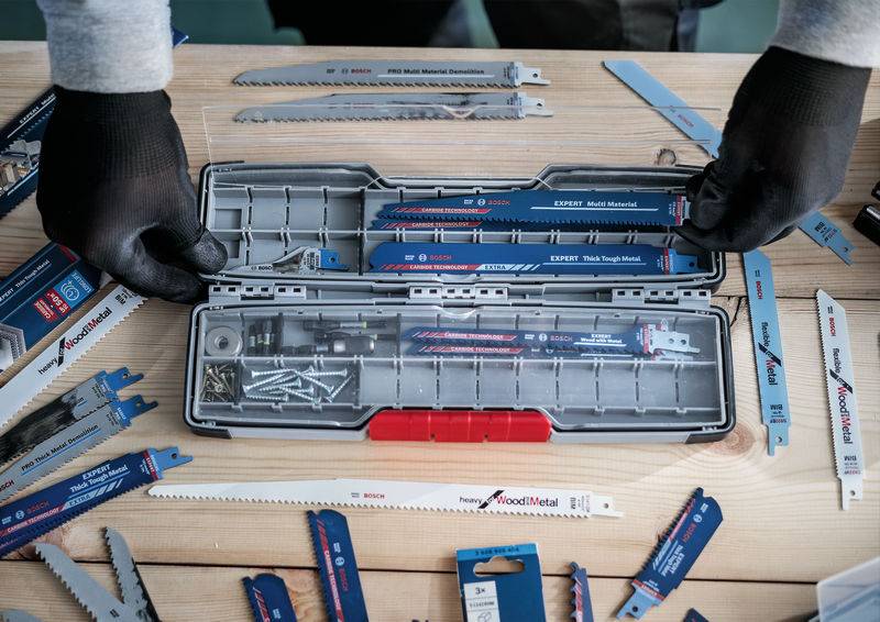 Una persona sta ordinando lame da sega in un contenitore degli attrezzi aperto su un tavolo di legno. Diverse lame da sega sono sparse tutt'intorno.
