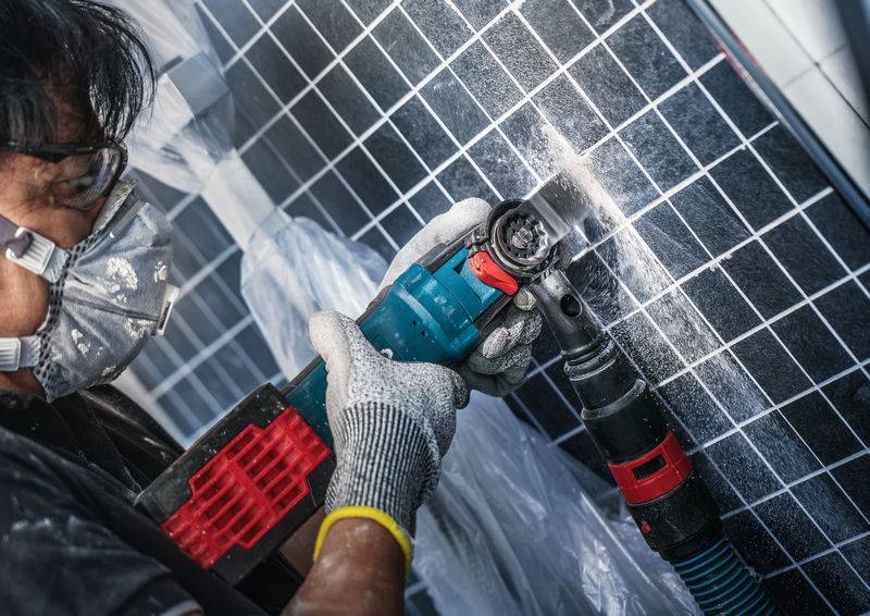 Persona con occhiali protettivi e maschera che taglia la superficie di un pannello solare con un utensile elettrico. Il posto di lavoro è coperto con un telo di plastica.