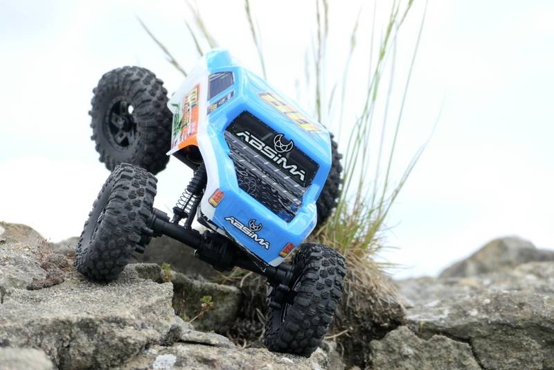 Un'auto radiocomandata con carrozzeria blu sta guidando su un terreno accidentato e rocce, con dell'erba visibile sullo sfondo.