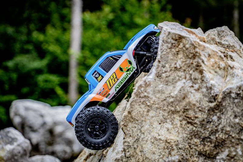 Un'auto giocattolo blu con il numero '112' scala un grande masso. Sullo sfondo si intravedono alberi sfocati.