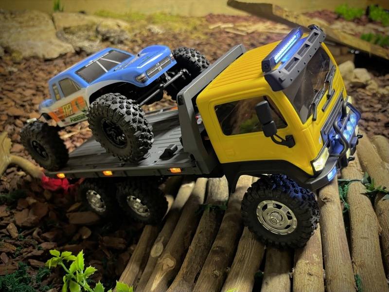 Due fuoristrada radiocomandati su un sentiero di legno in miniatura; il camion giallo trasporta un camion blu più piccolo sul suo pianale.