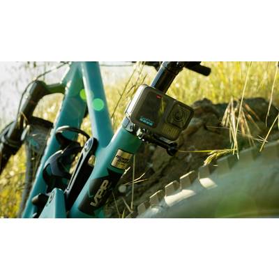 Una mountain bike blu con una action-camera montata è posta su un terreno roccioso, circondato da erba sotto la luce del sole.