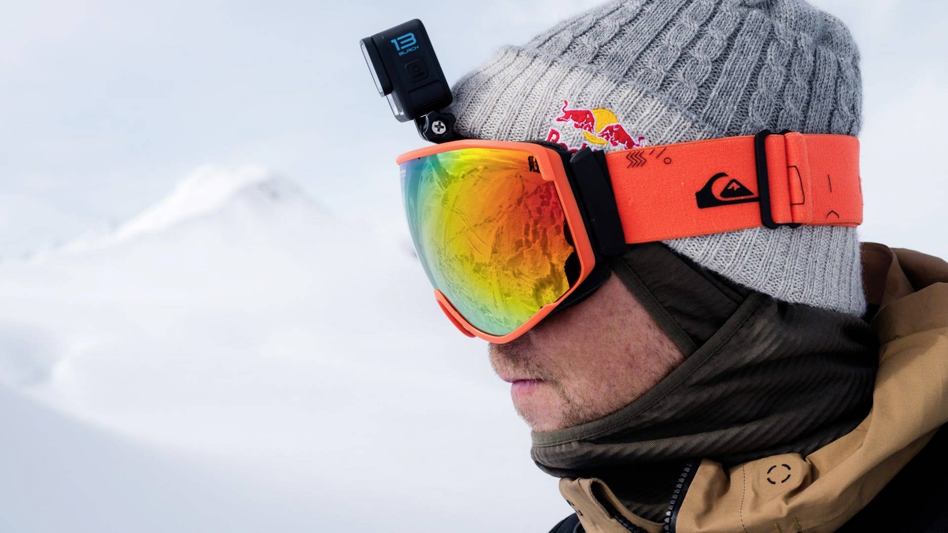 Una persona vestita con abiti invernali guarda verso destra. Indossa degli occhiali da sci arancioni e un cappello grigio con una macchina fotografica sopra. Montagne innevate sullo sfondo.