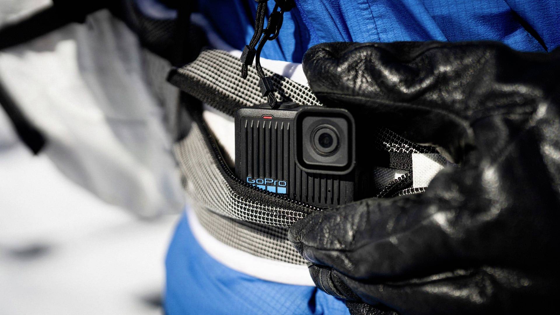 Una mano con un guanto nero tiene una piccola action camera fissata a una cintura. Sullo sfondo si vedono neve e abbigliamento blu.