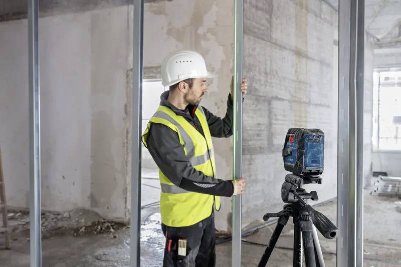 Un lavoratore edile che indossa un elmetto e un giubbotto riflettente sta regolando una trave verticale all'interno di una struttura parzialmente costruita, con un livello laser su un treppiede nelle vicinanze.