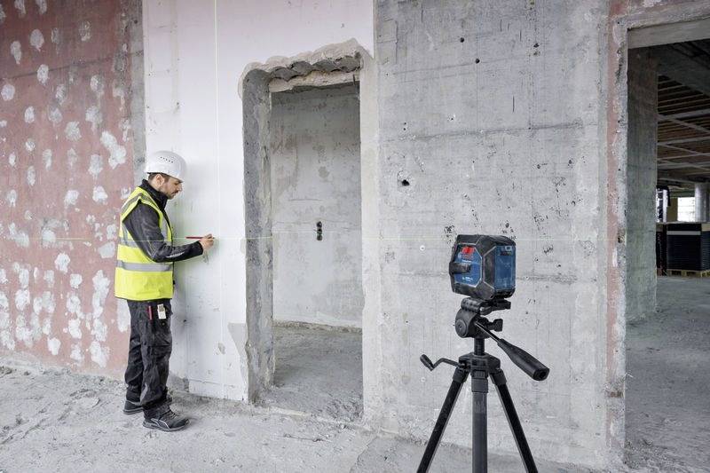 Un operaio edile che indossa un casco e un giubbotto di sicurezza misura un muro all'interno di un edificio in costruzione, utilizzando un livello laser montato su treppiede.
