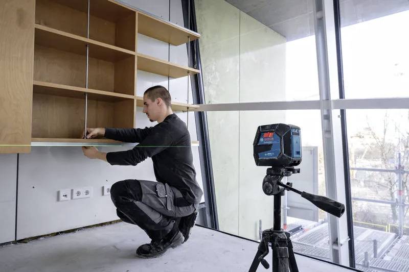 Una persona installa un mobiletto a parete in legno utilizzando un livello laser per l'allineamento in un edificio moderno, parzialmente costruito, con grandi finestre.
