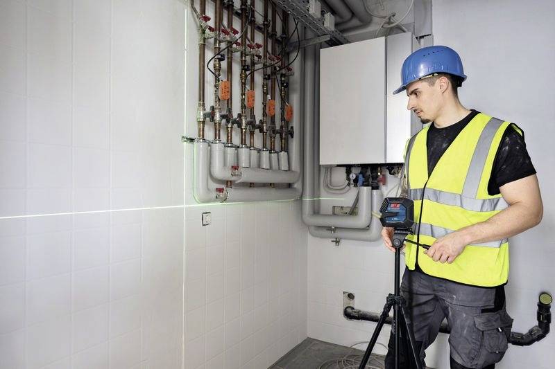 Un lavoratore con un giubbotto ad alta visibilità e un casco sta utilizzando uno strumento di misurazione laser in una sala macchine, allineandosi con tubi e pannelli.