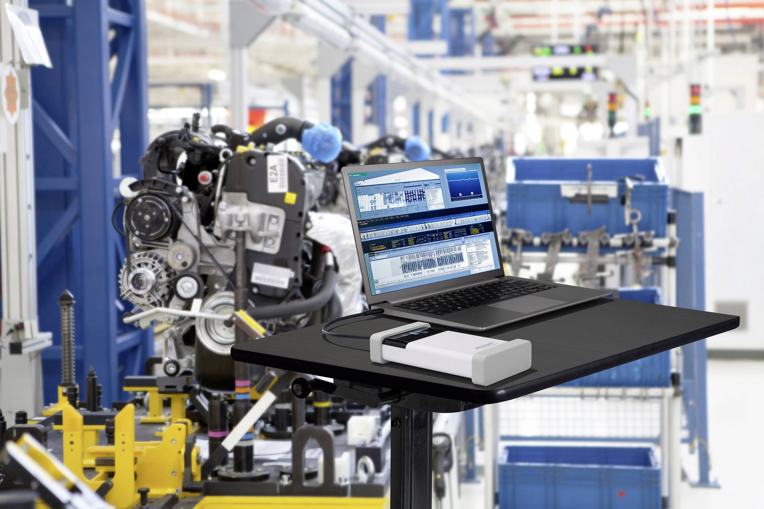 Un laptop è posizionato su un tavolo in una fabbrica. Lo schermo mostra un software di produzione. Sullo sfondo si vedono macchine e linee di produzione.