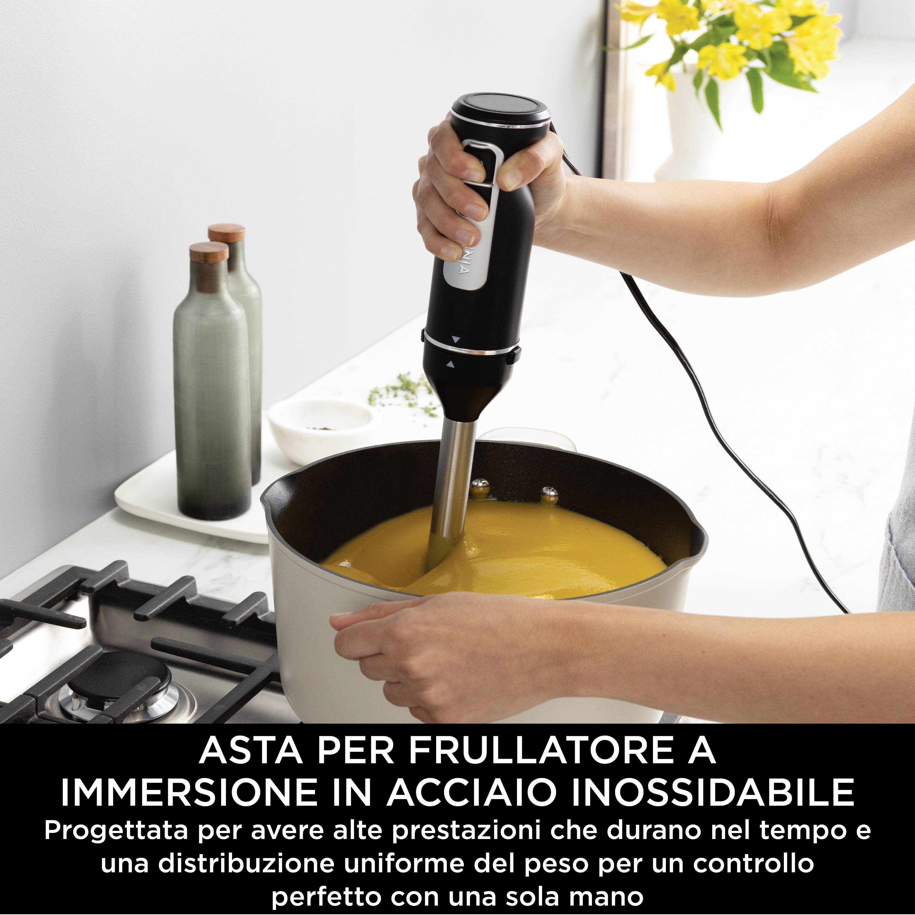 Una mano tiene un frullatore a immersione che viene utilizzato in una pentola con zuppa gialla su un fornello. Il testo sottostante descrive un prodotto.