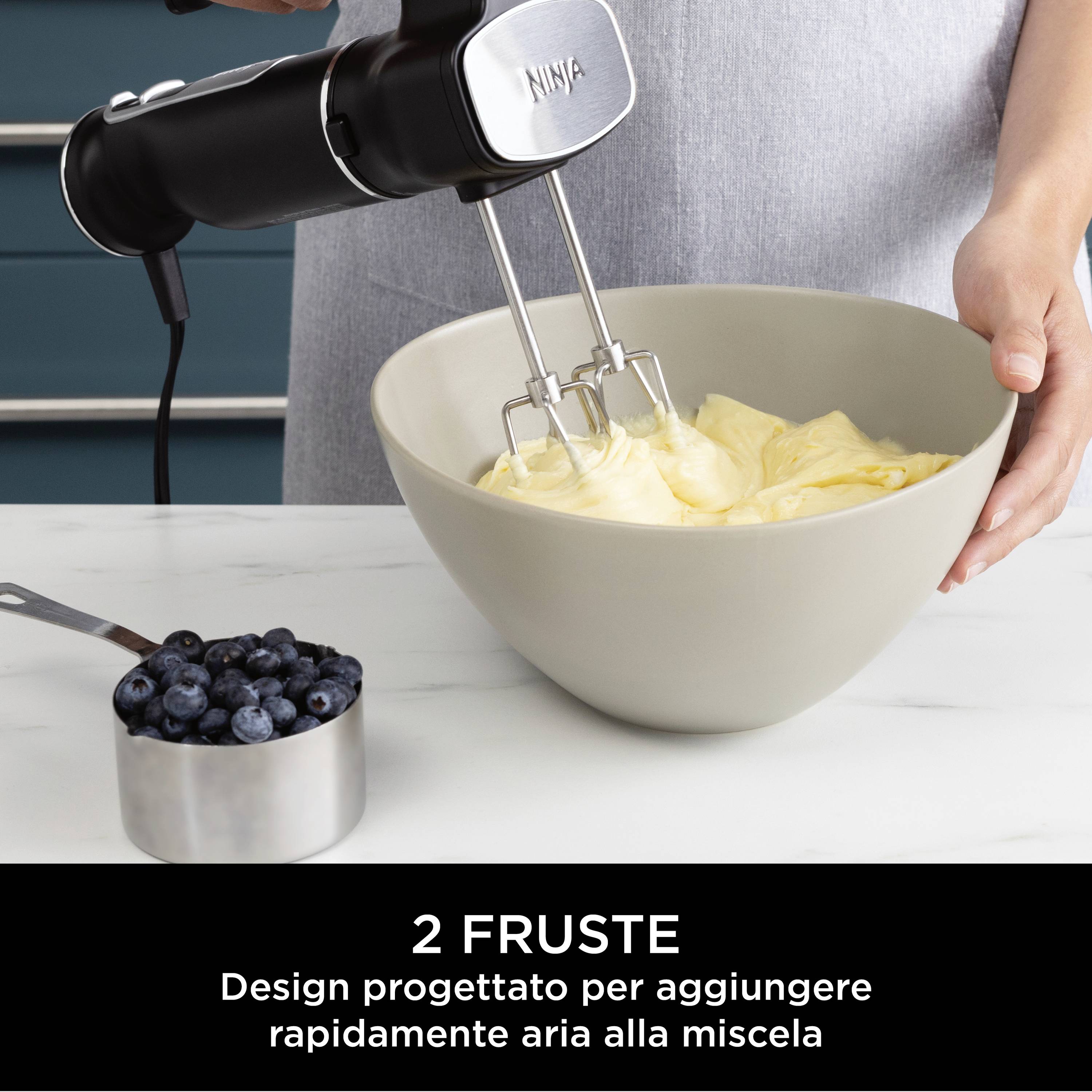 Mixer in azione: Una persona tiene un mixer nero sopra una ciotola con impasto. Accanto c'è una ciotola con mirtilli.
