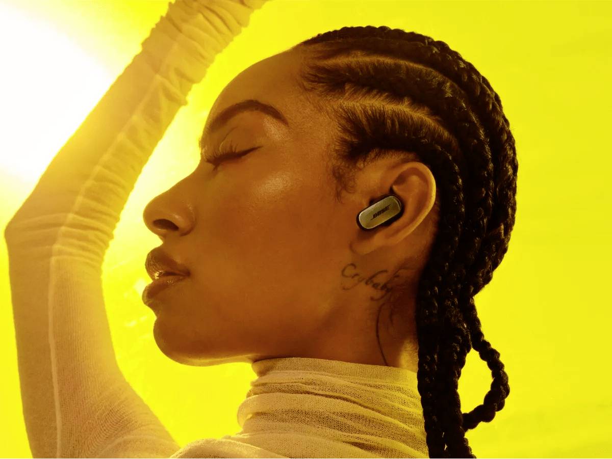 Una persona indossa auricolari wireless davanti a uno sfondo giallo, con gli occhi chiusi, in una posa rilassata. Trasmette una sensazione di tranquillità.