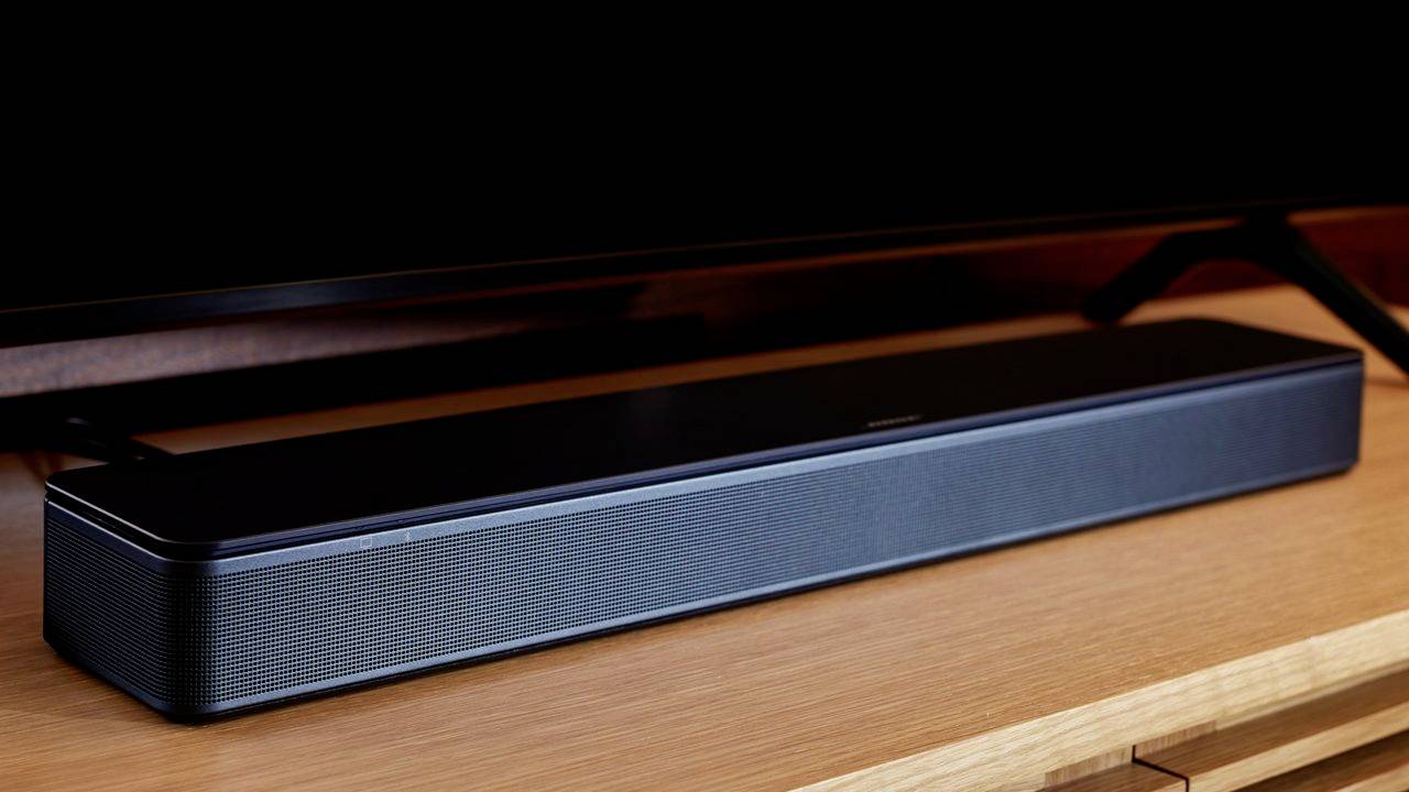 Soundbar nera su un tavolo di legno, posizionata davanti a un televisore a schermo piatto. Ideale per migliorare l'audio TV nelle sale soggiorno.
