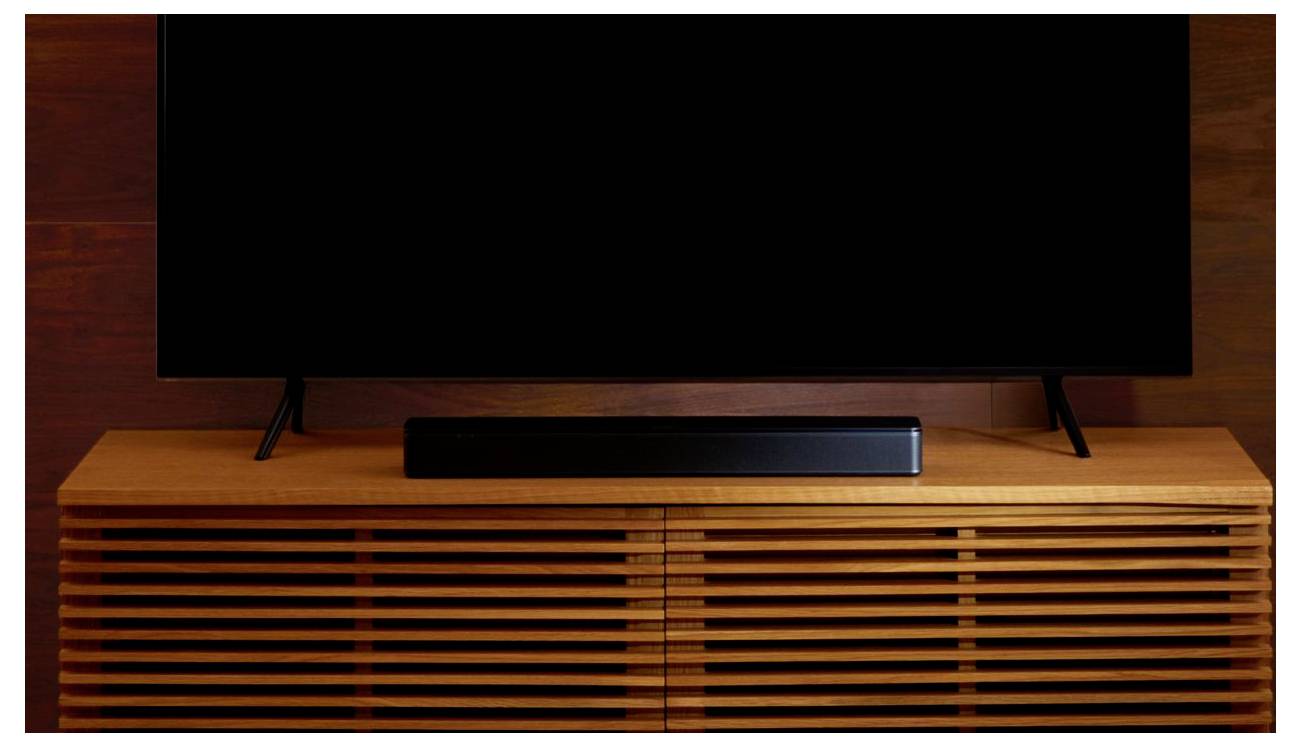 Una soundbar nera è posizionata su un mobile TV in legno. Sullo sfondo, è visibile il bordo inferiore di un televisore a schermo piatto.