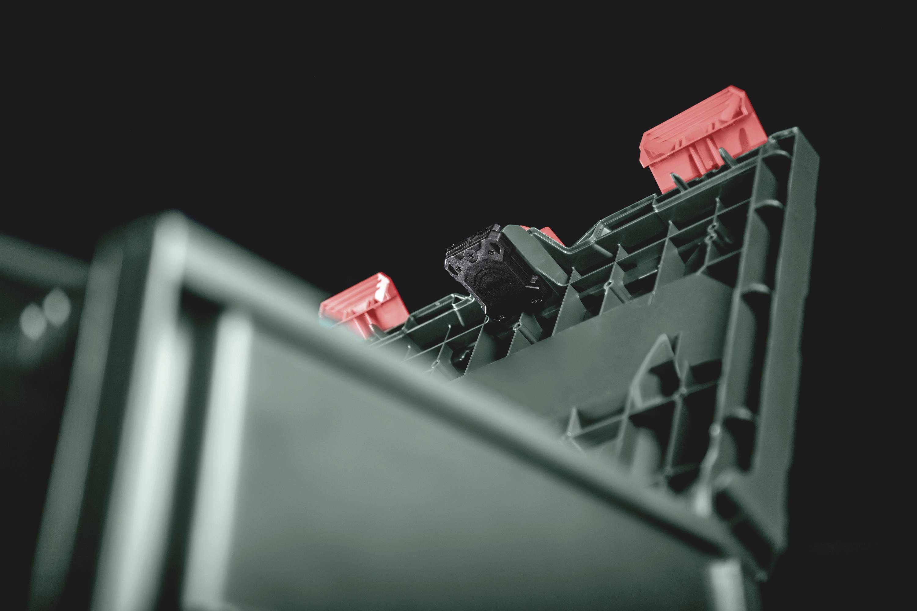 Primo piano di un contenitore nero in plastica con chiusure rosse, su sfondo scuro. La struttura del contenitore è ben visibile.