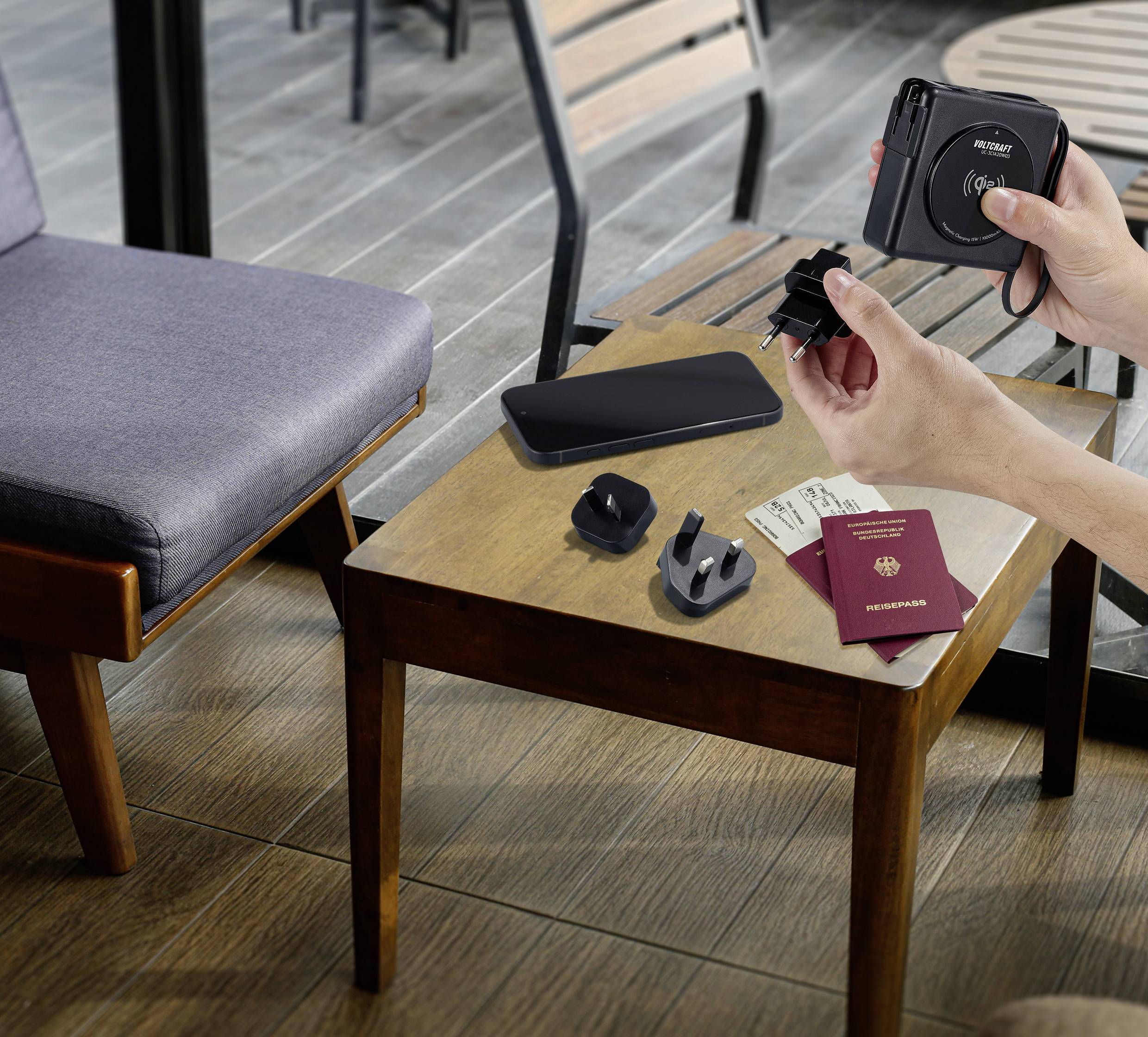 Una persona mostra un adattatore universale da viaggio sopra un tavolo con uno smartphone, diversi attacchi per adattatori e un passaporto.