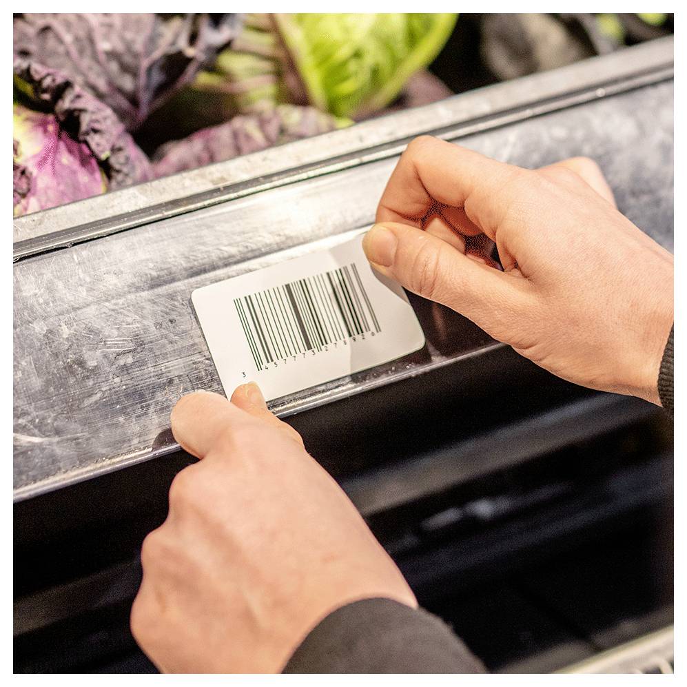 'Primo piano di mani che scansionano un'etichetta con codice a barre su uno scaffale in un supermercato, con verdure visibili sullo sfondo.'
