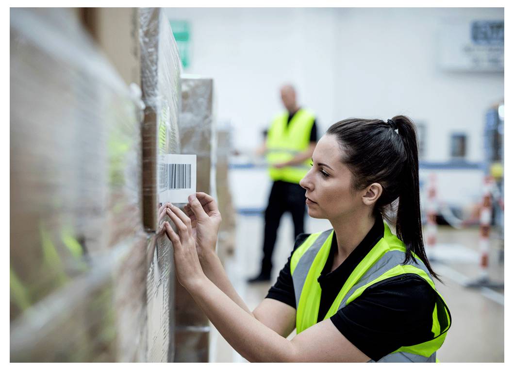 Un lavoratore con un giubbotto alta visibilità, concentrato nello scansionare un codice a barre su un pallet in un magazzino, mentre un altro lavoratore è in piedi sullo sfondo.