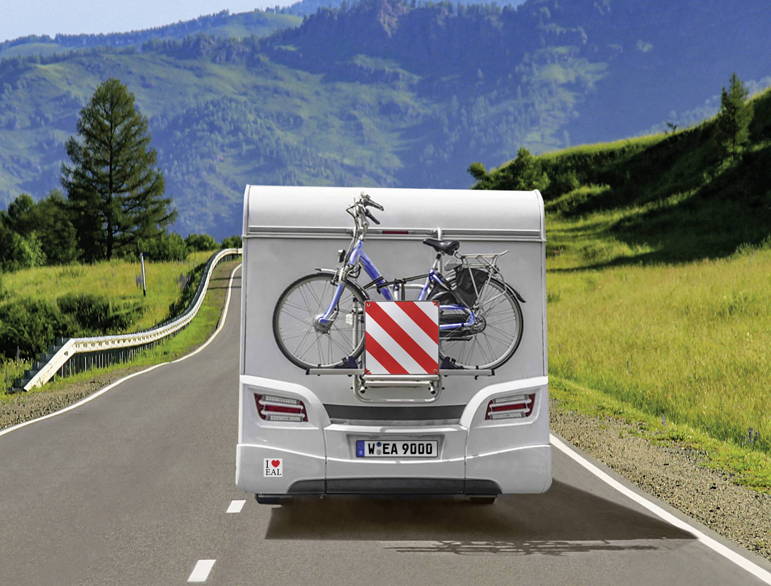 Un camper percorre una strada statale. Sul retro è montata una bicicletta contrassegnata da un'avvertenza a strisce rosse e bianche. Sullo sfondo si vedono colline verdi.