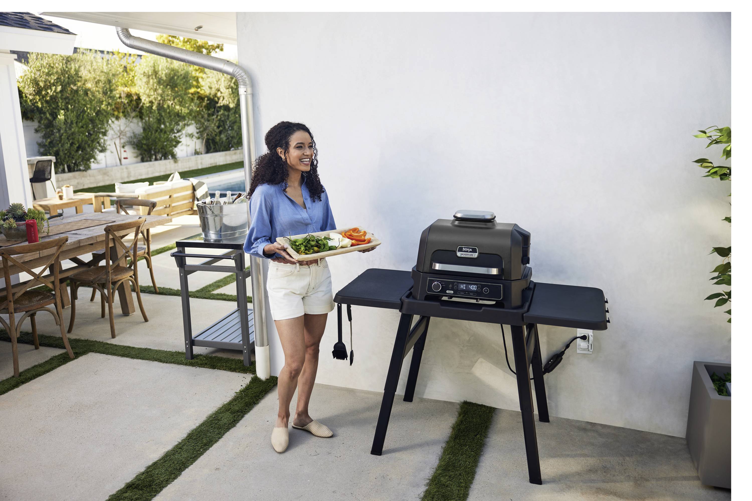 Una donna è in piedi in un giardino accanto a un barbecue e tiene in mano un piatto di verdure. Sullo sfondo si intravede un tavolo esterno.