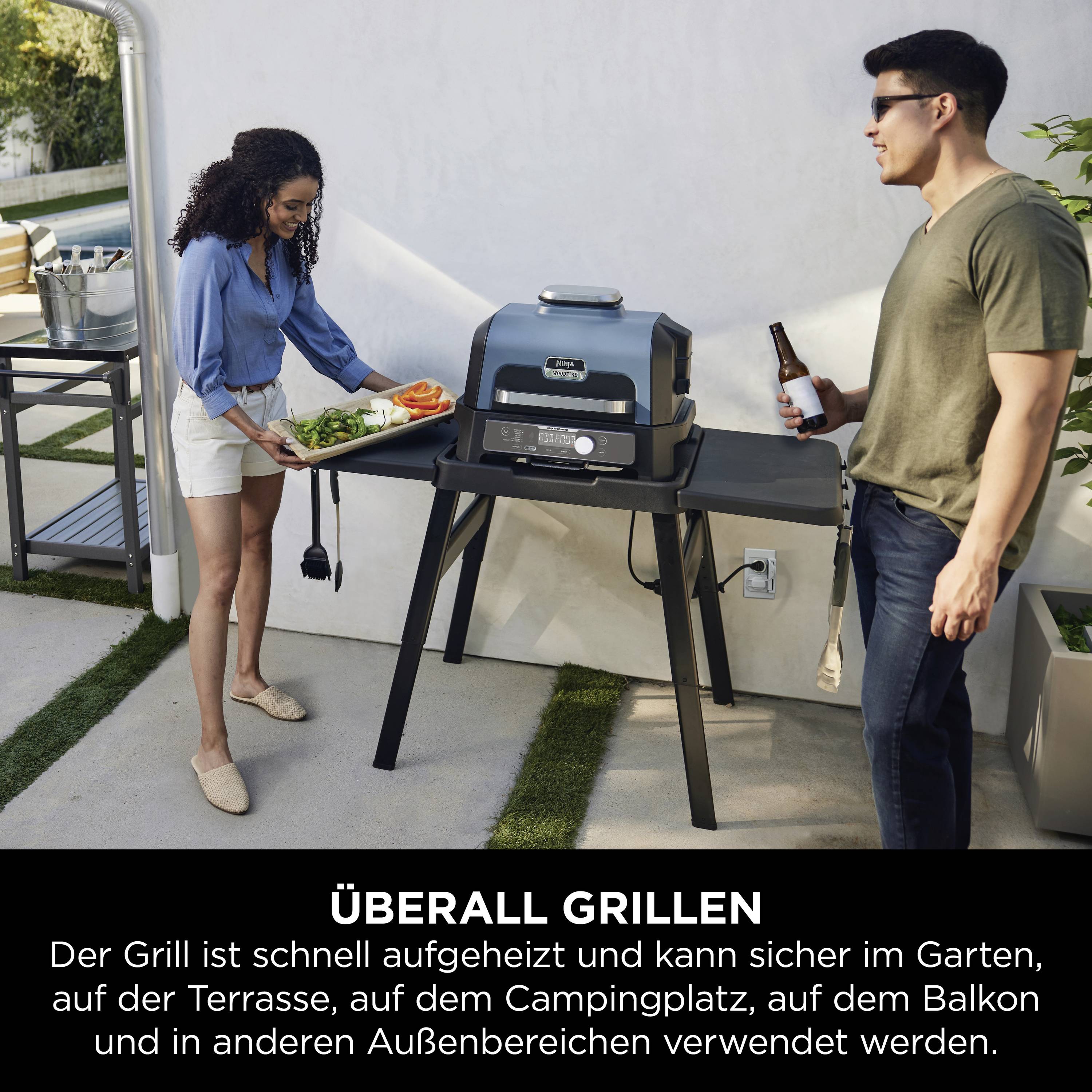 Una donna porta verdure a un barbecue, un uomo tiene in mano una bevanda. Il barbecue è nel giardino accanto a un tavolo con utensili.
