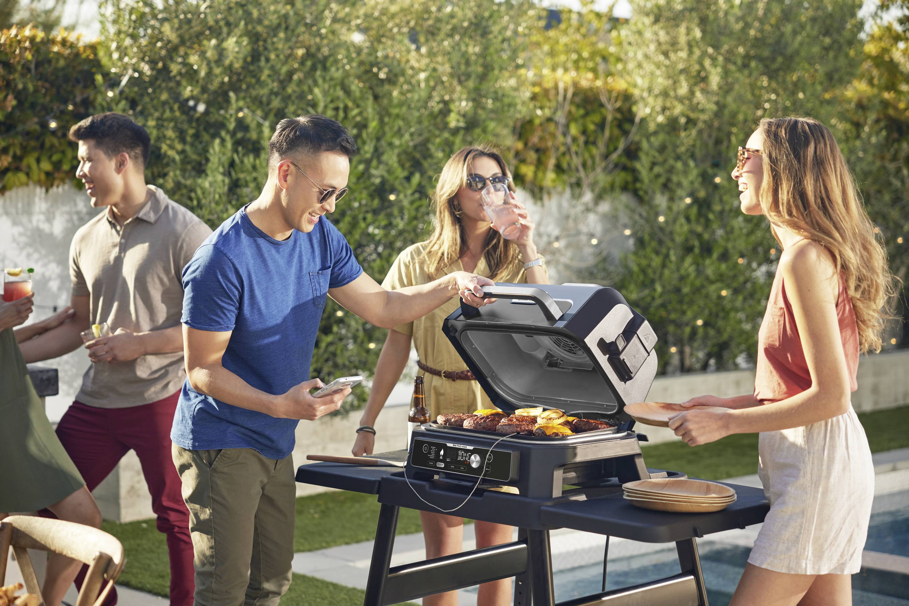 Gruppo di persone che fa un barbecue in giardino. Diversi adulti sono radunati intorno a un barbecue, mentre due persone stanno mettendo delle bistecche sulla griglia. Tutti sembrano rilassati e allegri.