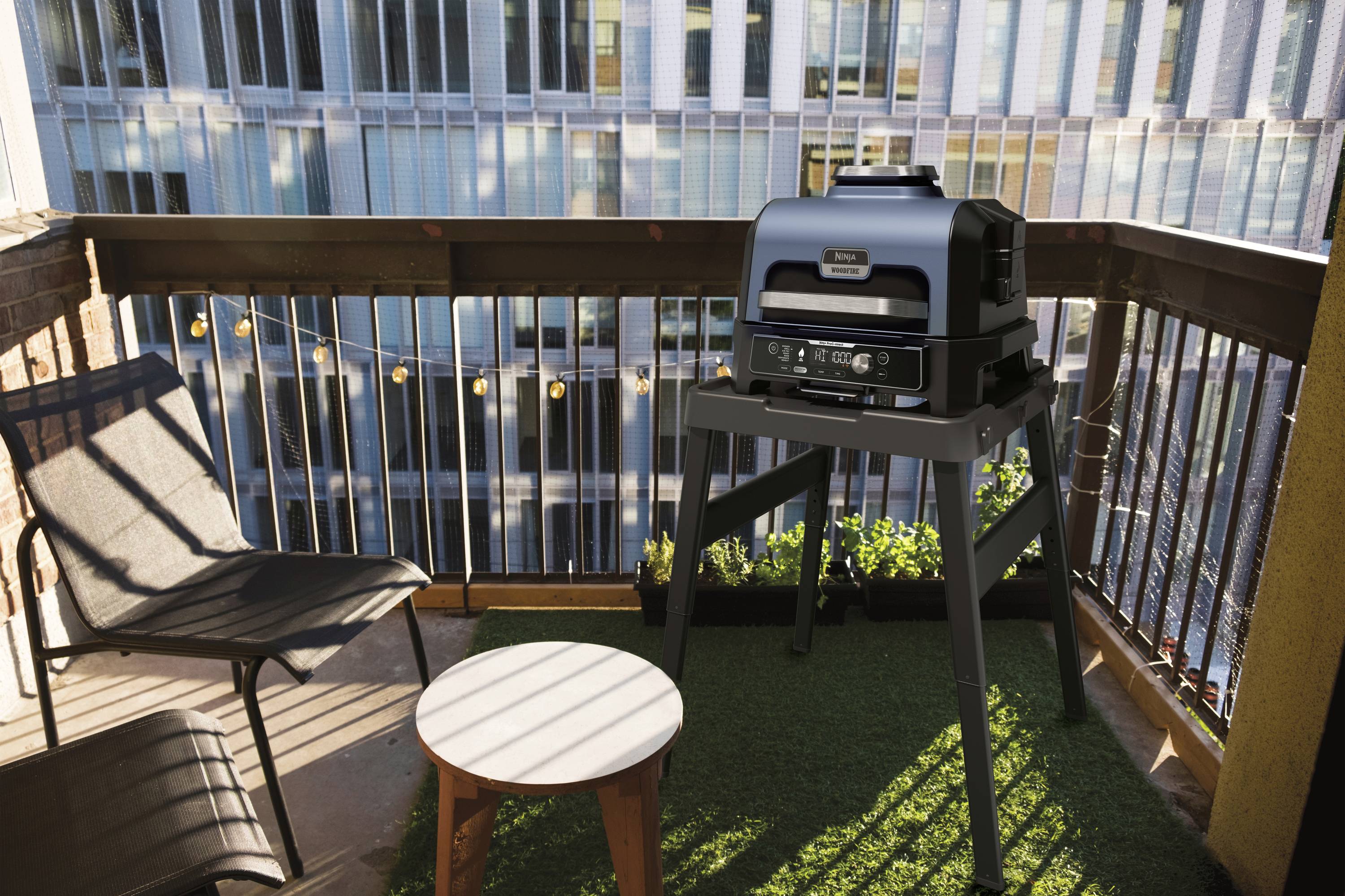Un barbecue moderno è posizionato su un balcone con pavimento in legno e tappeto erboso sintetico. Accanto c'è un piccolo tavolo e una sedia. Sullo sfondo si vede un edificio.