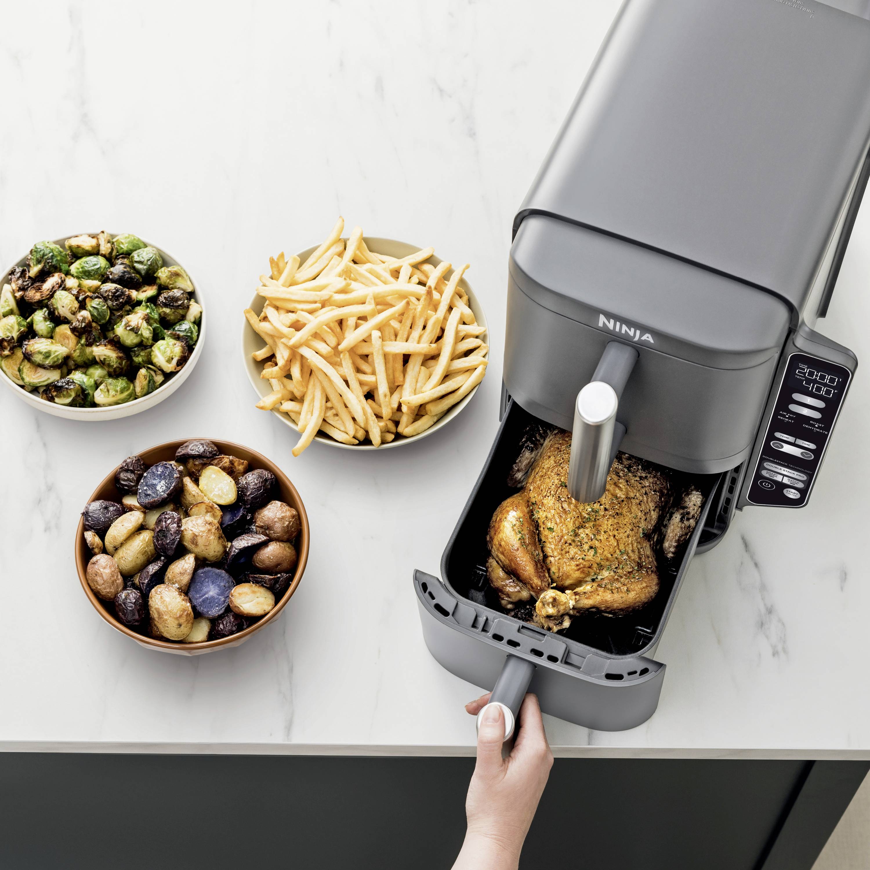 Una mano sta azionando una friggitrice ad aria con un pollo fritto. Accanto, un piatto con patatine fritte, cavoletti di Bruxelles e patate.