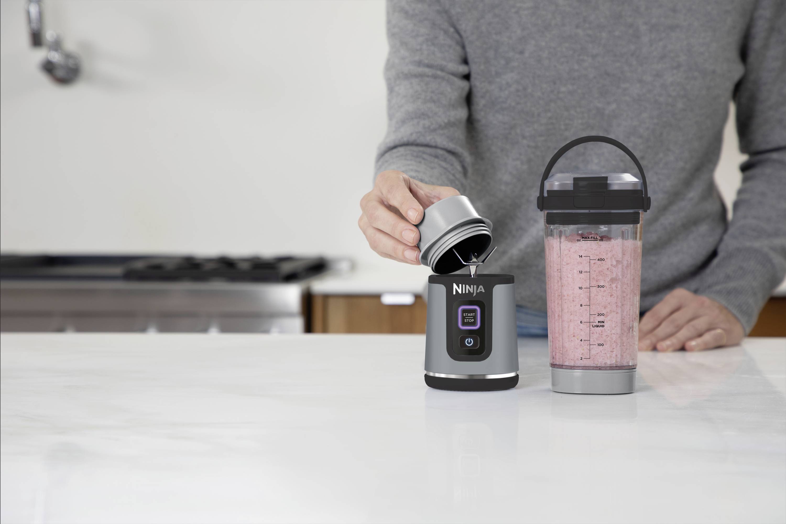Una persona sta preparando frullati con un frullatore a immersione. Il mixer è posizionato su un piano di lavoro bianco in cucina, accanto a un bicchiere riempito.
