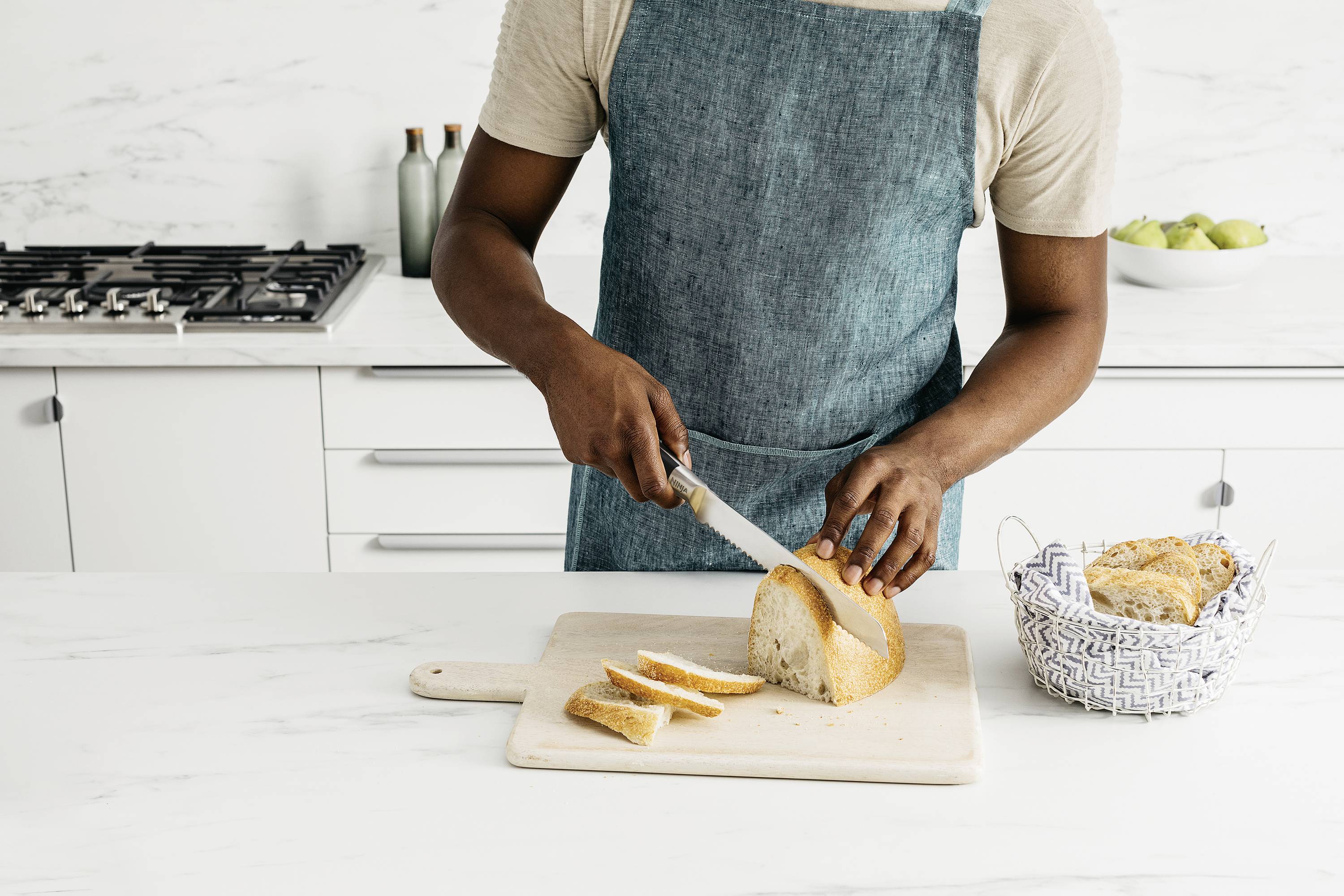 Una persona sta tagliando il pane a fette su un tagliere di legno. Un cestino del pane è posizionato accanto. La cucina è arredata in stile moderno.