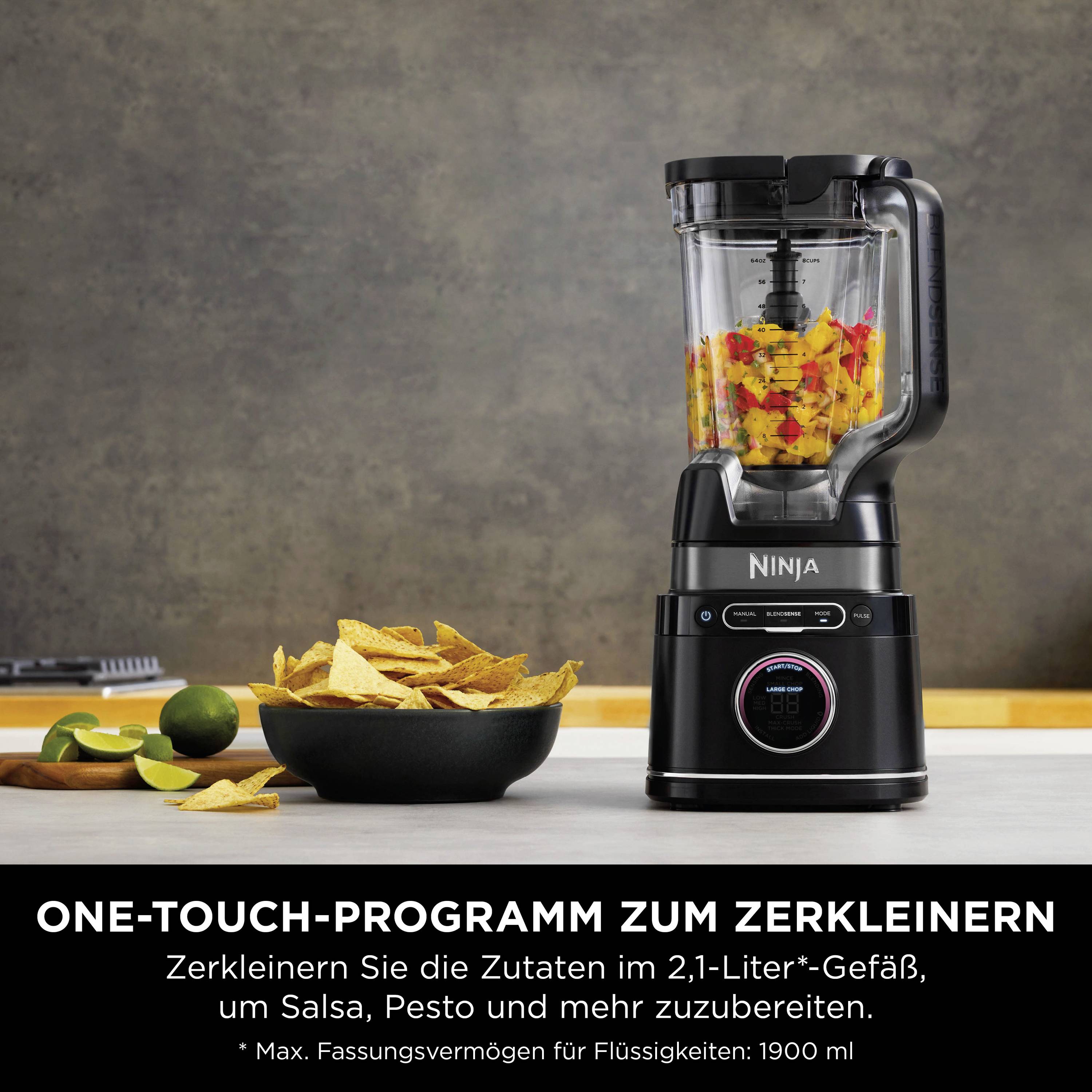 Un frullatore con un bicchiere, riempito con ingredienti tritati, è posizionato su un piano di lavoro. Accanto ci sono ciotole con chips e lime.