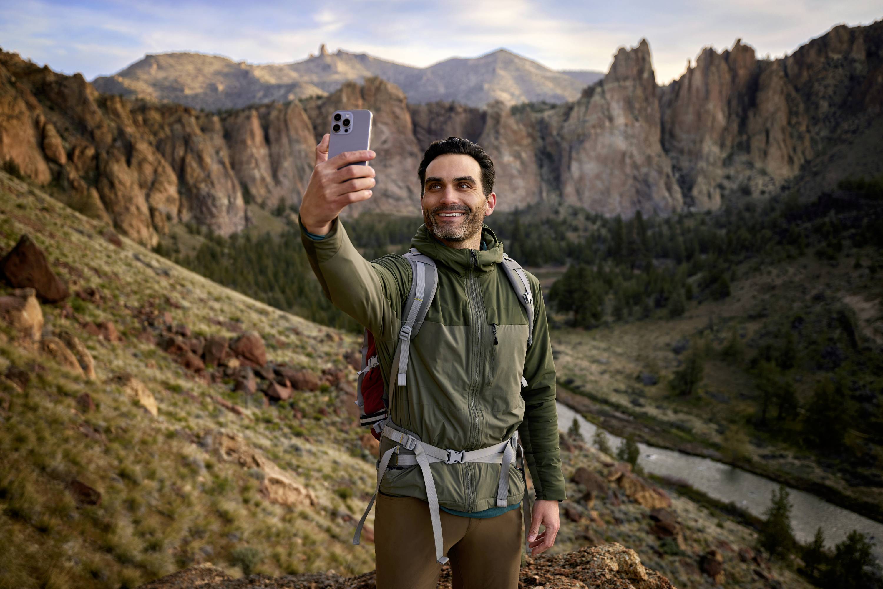 Una persona su un sentiero escursionistico in un paesaggio montuoso si scatta un selfie. Sullo sfondo si vedono rocce e un fiume.