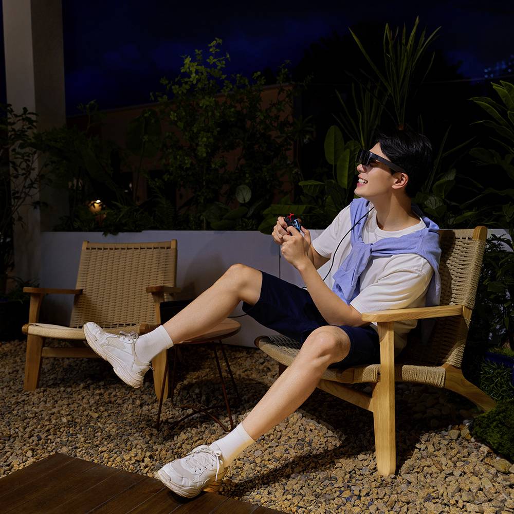 Un uomo seduto di notte, sorridente, in una poltrona in giardino, indossa occhiali da sole scuri e tiene in mano una piccola confezione rossa.