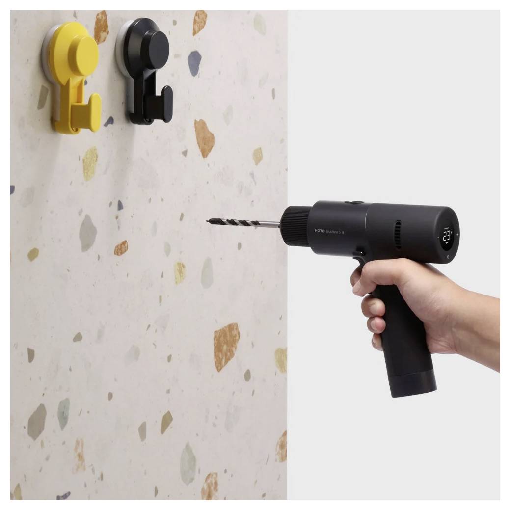 Una persona utilizza un trapano cordless nero su una parete in terrazzo con due ganci colorati. L'immagine dimostra un'attività di miglioramento domestico.