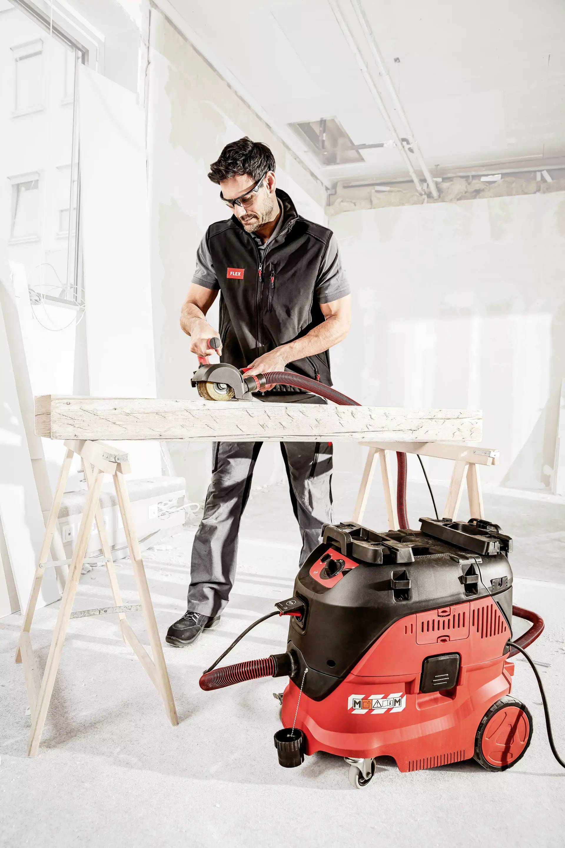 Un artigiano taglia legno con una sega in un cantiere. In primo piano è posizionato un aspiratore industriale rosso.
