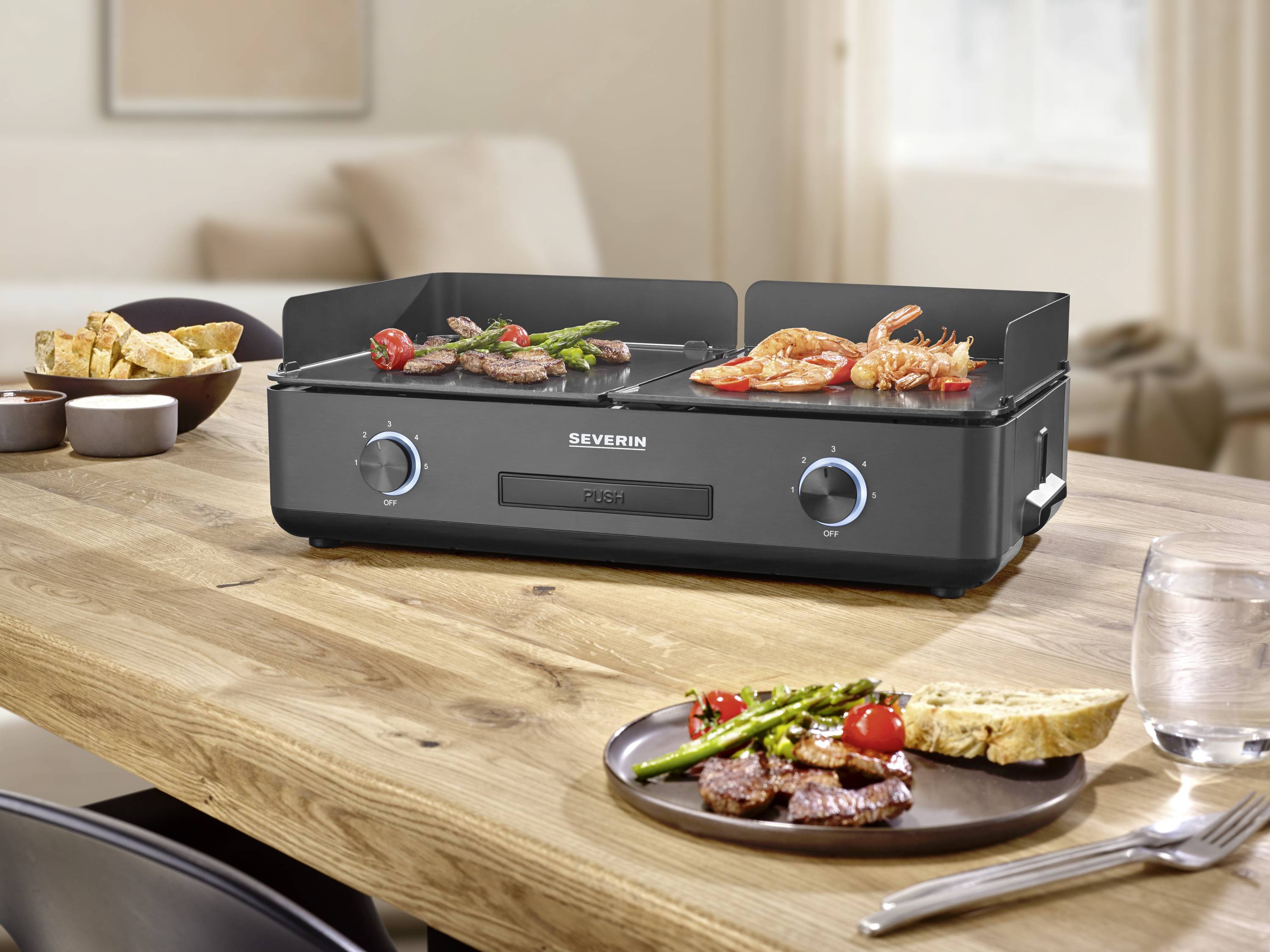Un grill da tavolo nero Severin con verdure e carne grigliate, circondato da piatti con del cibo, è posizionato su un tavolo di legno in una cucina.
