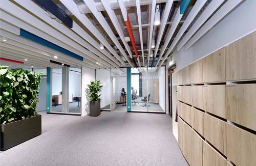 Un ufficio moderno con pareti di vetro, piante e mobili in legno. Un'illuminazione luminosa e un design aperto creano un'atmosfera accogliente.