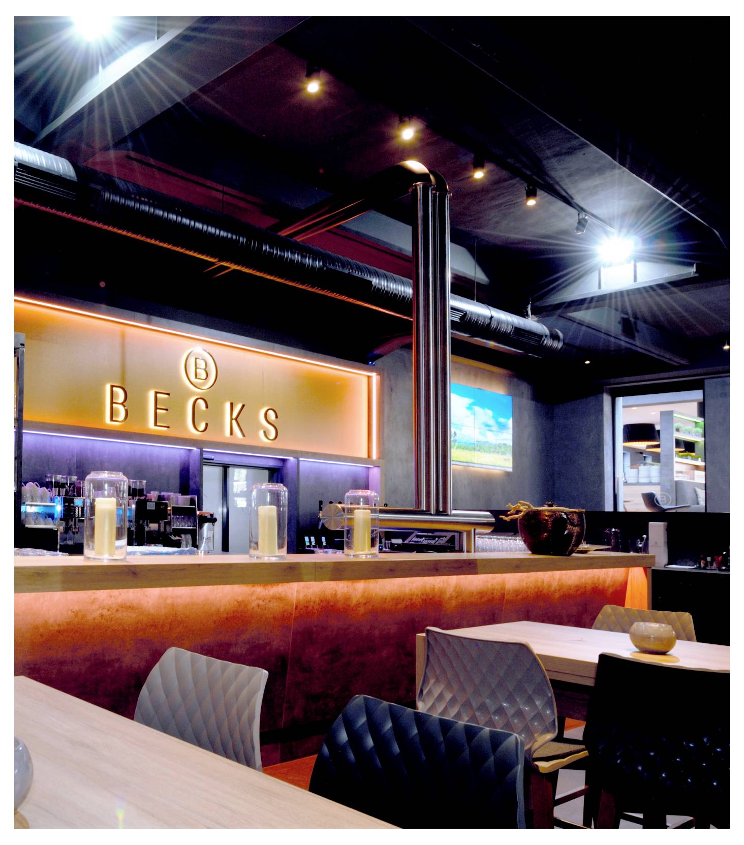 Interno di un bar o ristorante elegante con l'insegna illuminata "BECKS" dietro il bancone, posti a sedere in primo piano e un'illuminazione calda e soffusa.