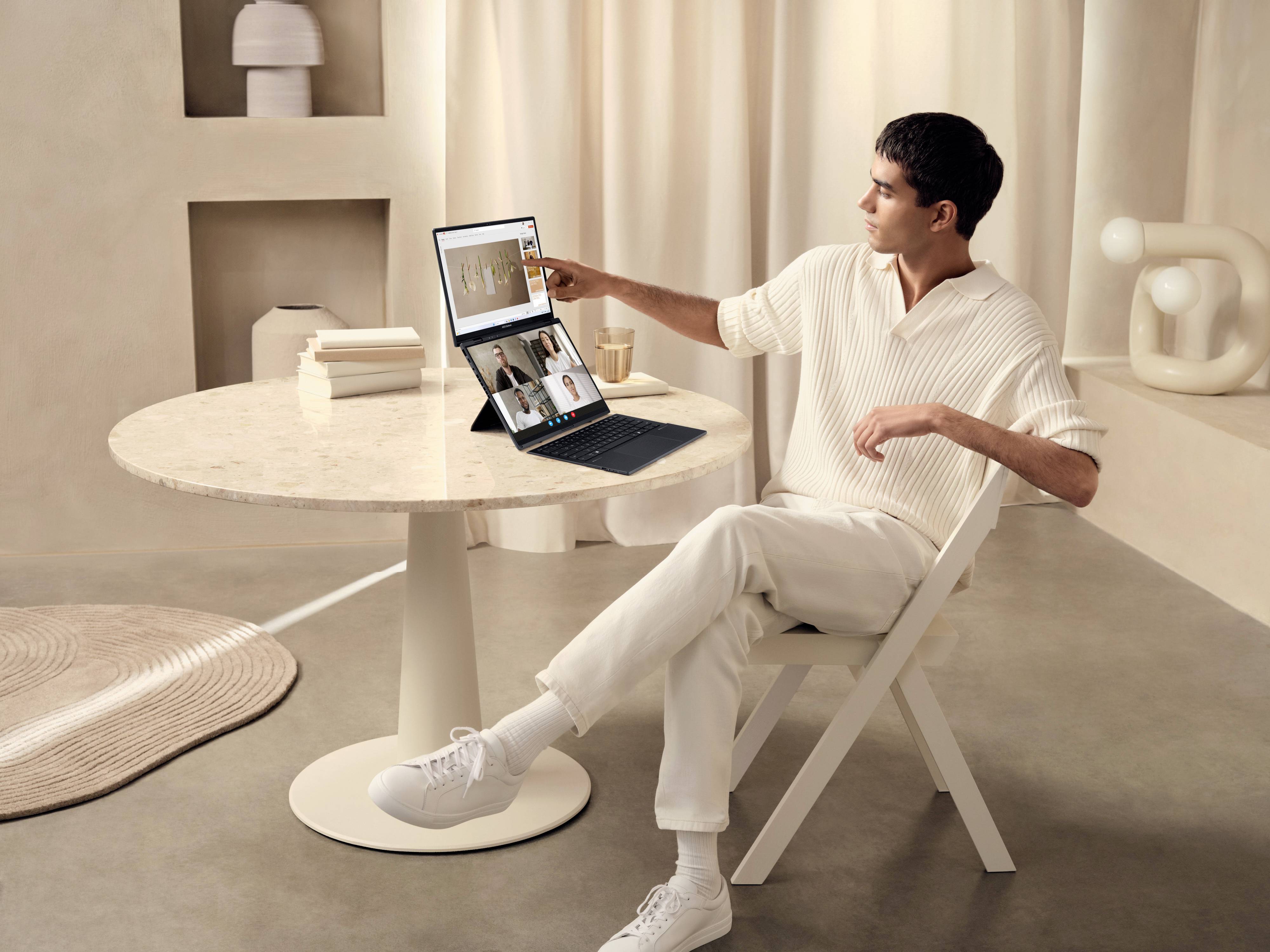 Un uomo è seduto a un tavolo e utilizza un laptop con un secondo schermo. Sullo schermo è visibile una videoconferenza. La stanza ha un arredamento minimale e luminoso.