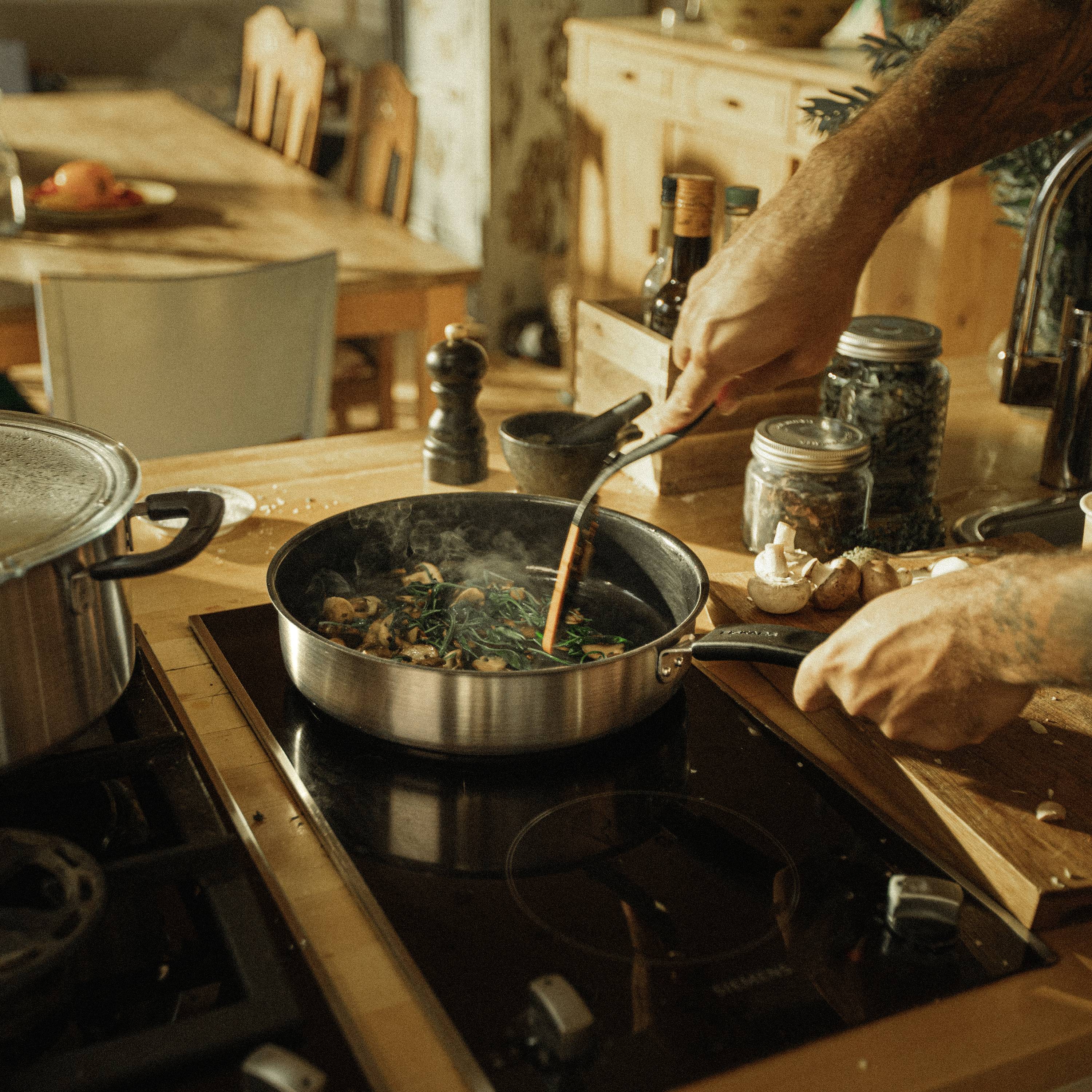 Una persona sta cucinando un pasto in una padella su un fornello, circondato da vari utensili da cucina e spezie. Un tavolo di legno fa da sfondo.