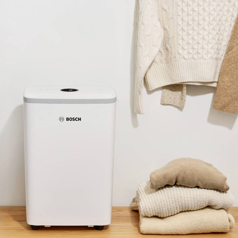 Un purificatore d'aria di Bosch è posizionato sul pavimento accanto a una pila di asciugamani beige e un cardigan appeso.