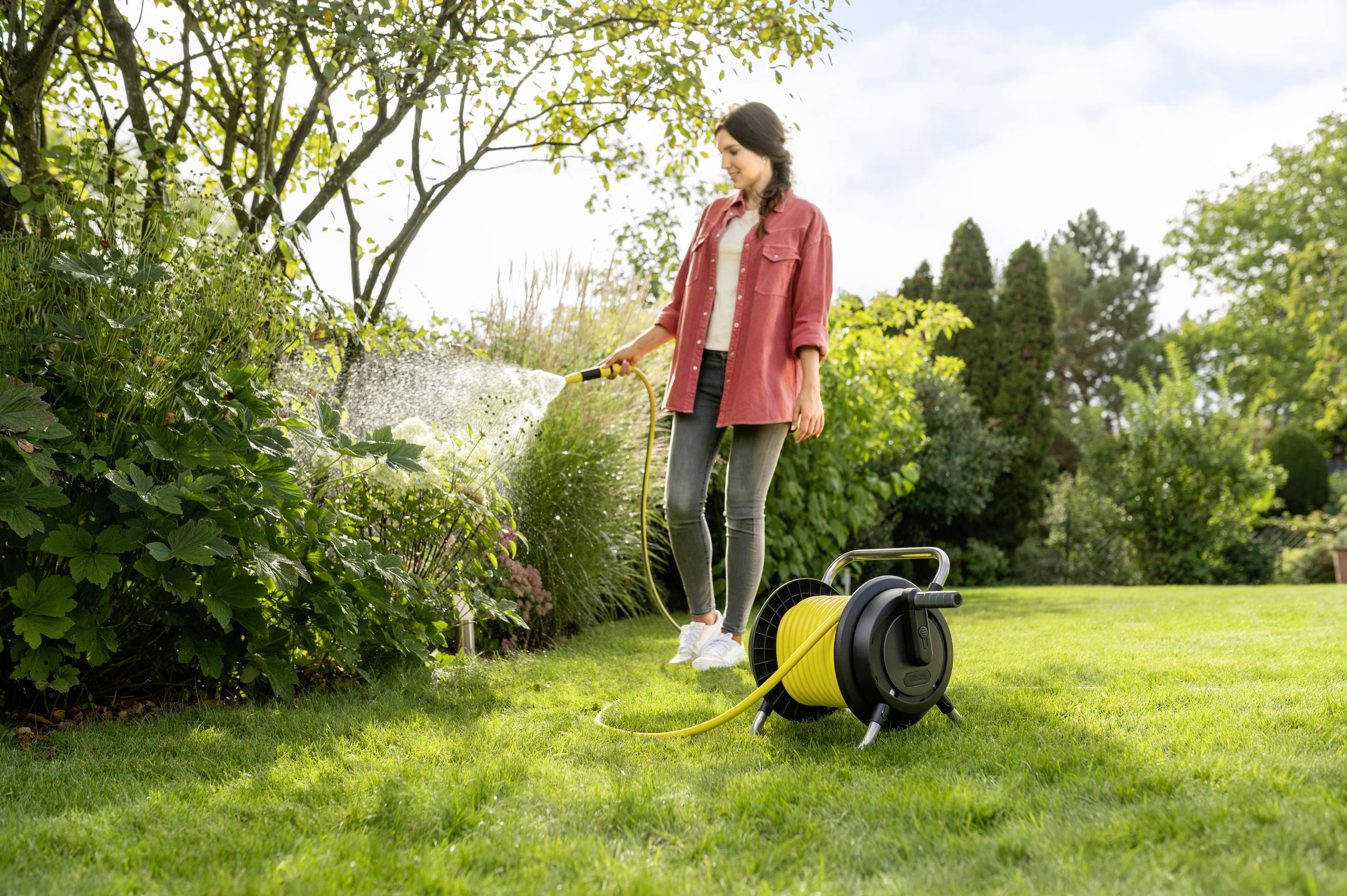 Una donna con una giacca rossa sta innaffiando i fiori nel giardino con un tubo dell'acqua. Un avvolgitubo è posizionato accanto a lei sull'erba.