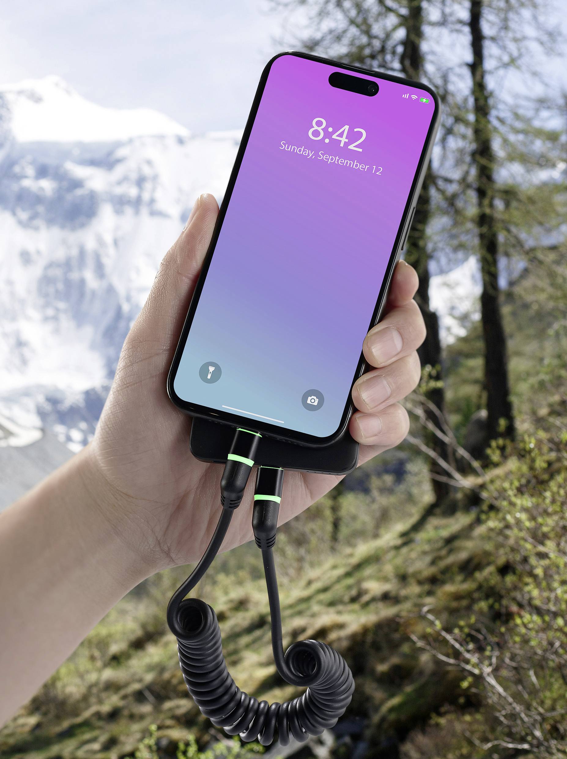 Una mano tiene uno smartphone con uno schermo viola-blu che mostra '8:42 Domenica, 12 settembre'. Sullo sfondo ci sono montagne e alberi.