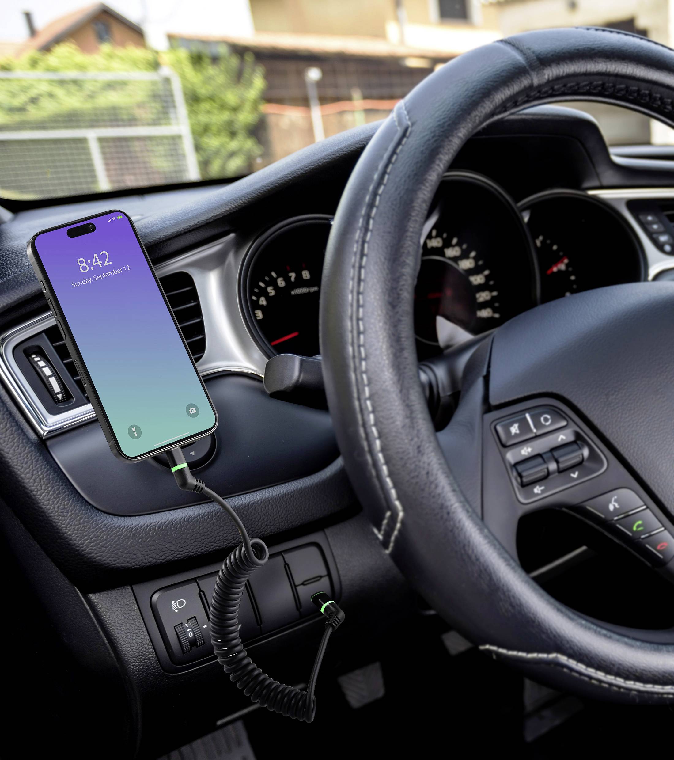 Smartphone collegato con cavo, agganciato al supporto del cruscotto in auto. Ora 8:42, sole all'esterno. Mostra connessione e utilizzo durante la guida.