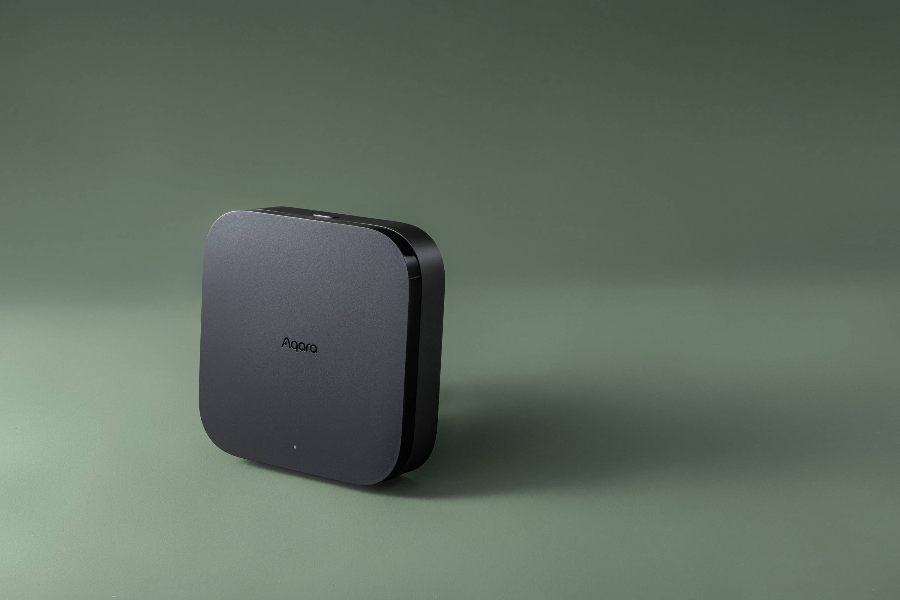 Un dispositivo quadrato nero di 'Aqara' è posizionato su uno sfondo verde, presumibilmente un prodotto tecnologico.