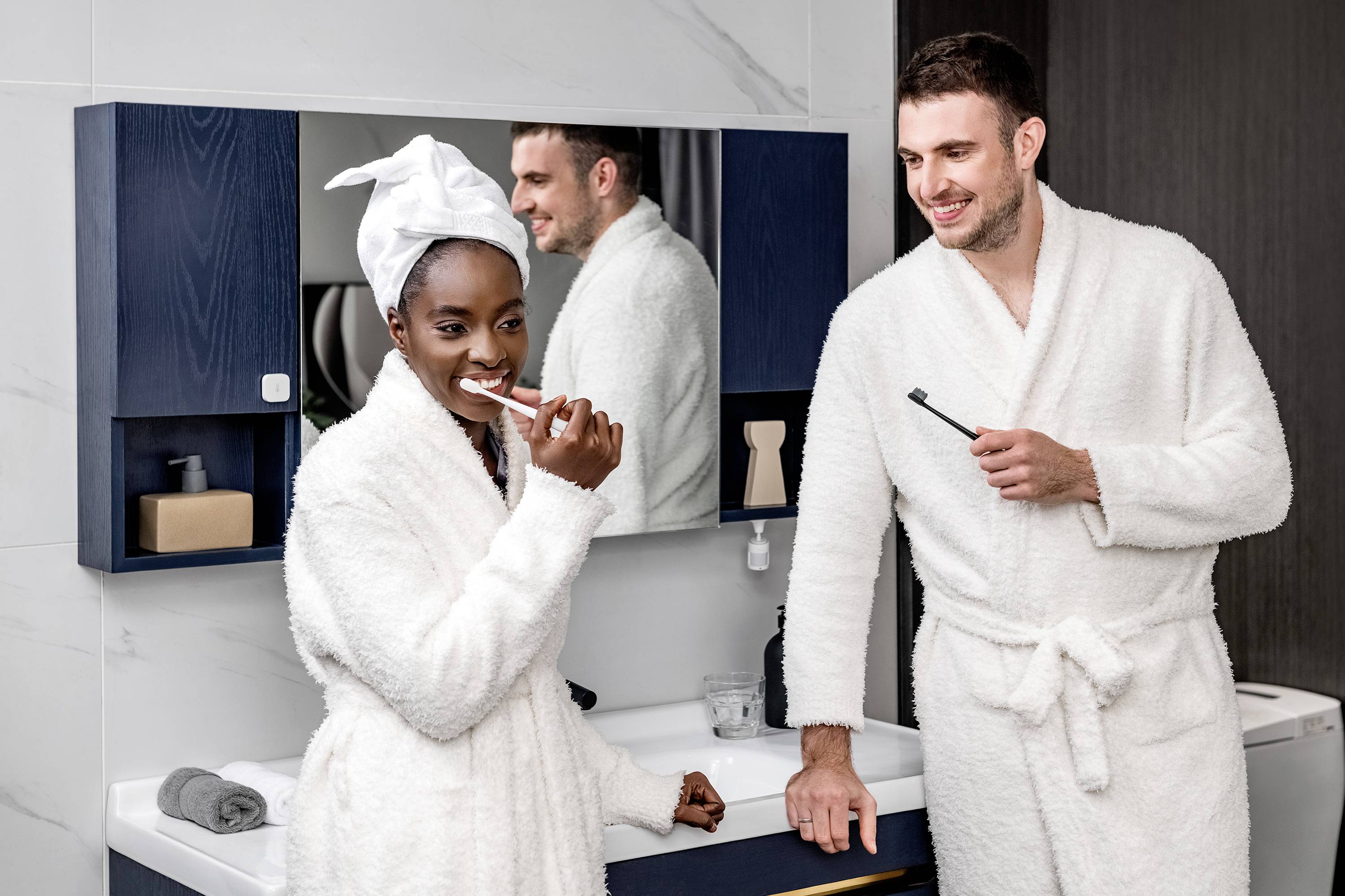 Una donna e un uomo in accappatoio stanno sorridendo in bagno. La donna si sta lavando i denti, mentre l'uomo la guarda.